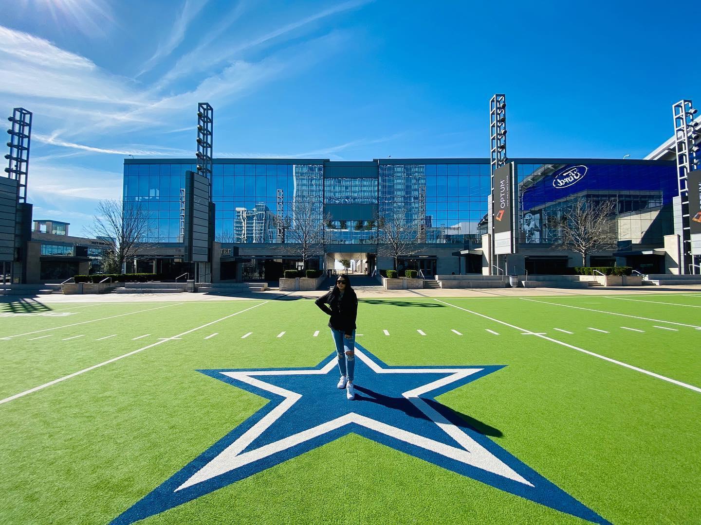 A woman poses on a large blue star on the Dallas Cowboys field on a sunny day.