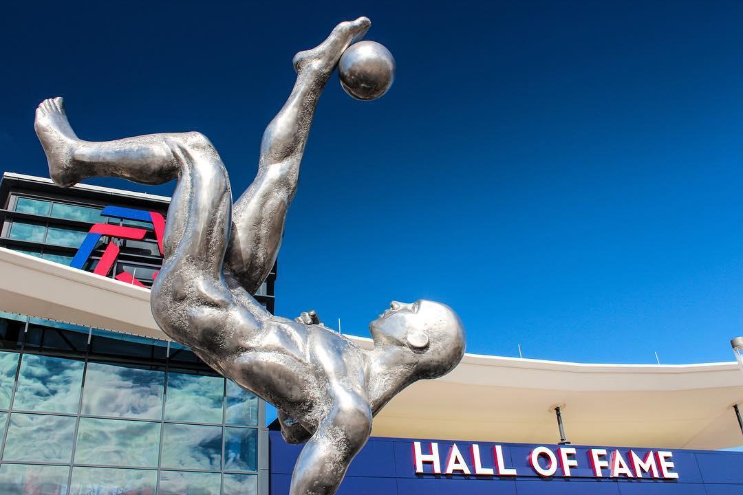 A large silver statue of a man kicking a soccer ball in front of the Soccer Hall of Fame.
