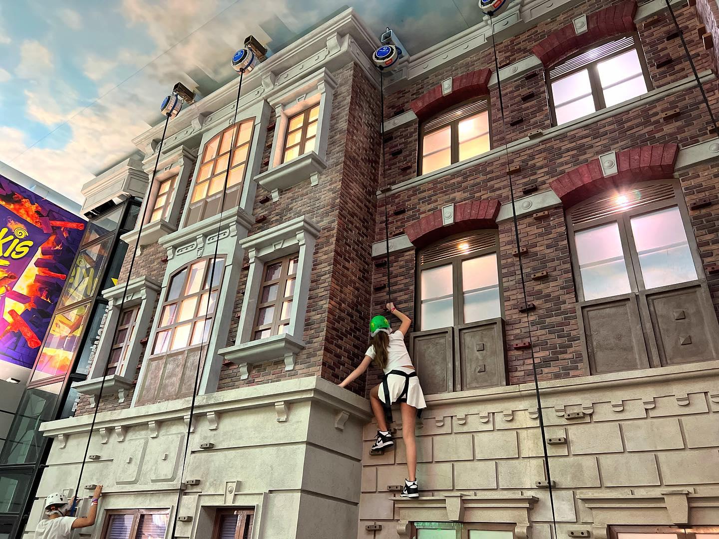 A young girl in rock climbing protective equipment climbs a wall designed to look like the exterior of a brick building.