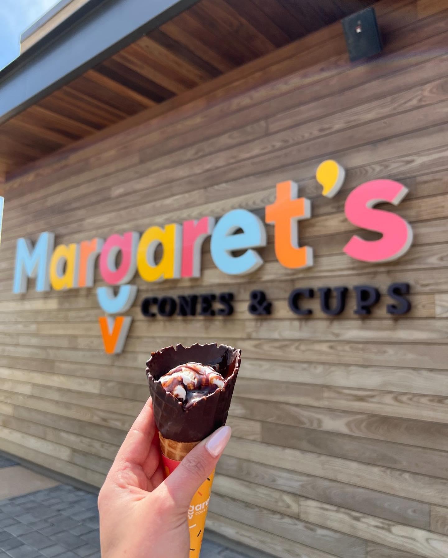 A manicured hand holds up an ice cream cone with cupcake inside in front of a colorful sign that says Margaret's.