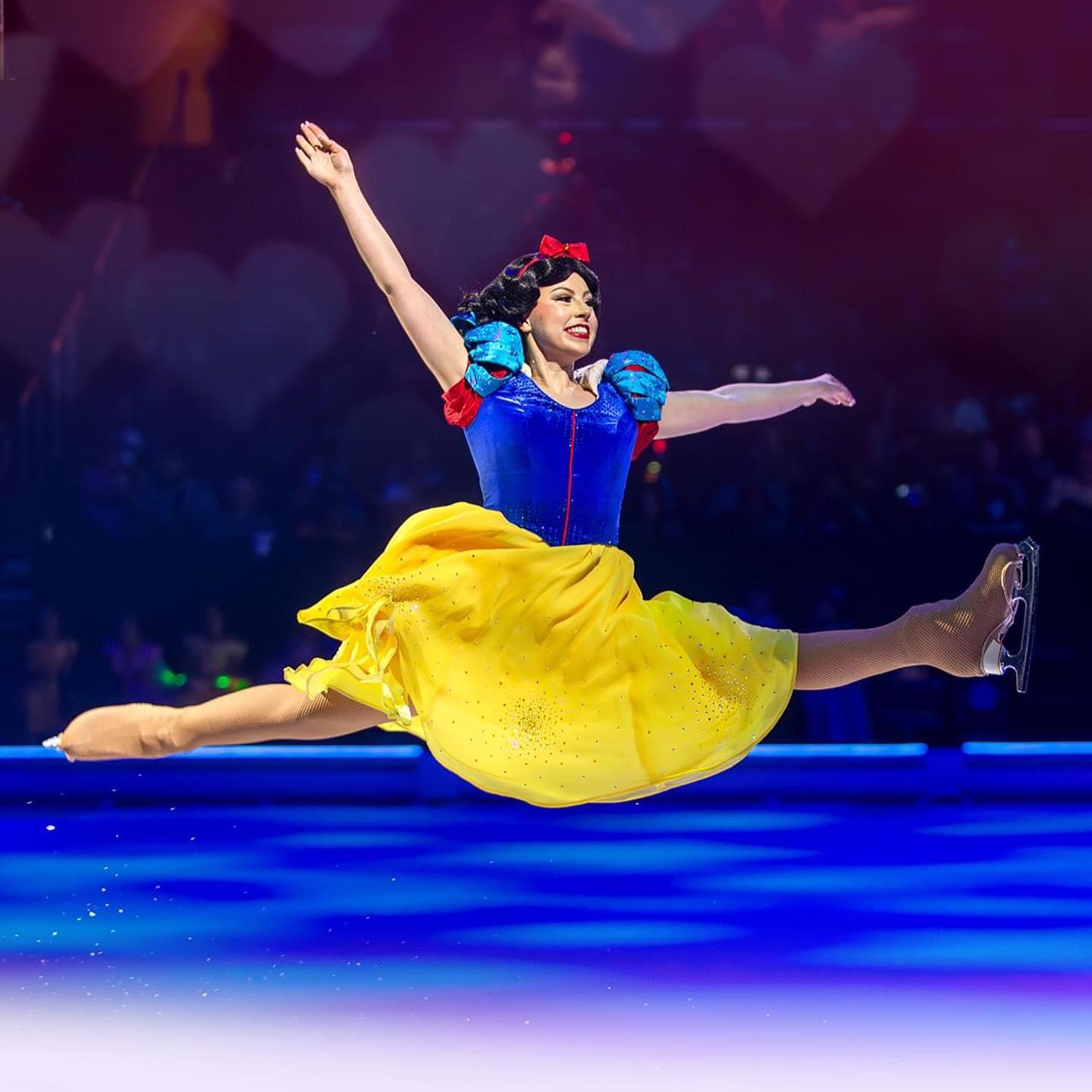 A ballerina jumps dramatically on stage while dressed as Snow White, with a bright yellow skirt and deep blue top.