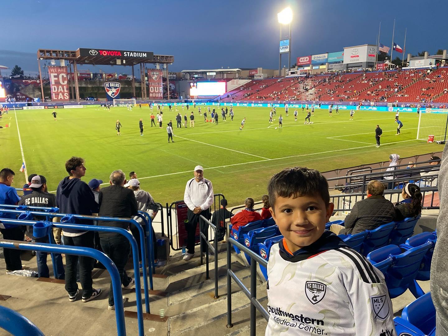 A young boy smiles for a photo with a baseball field in the background.