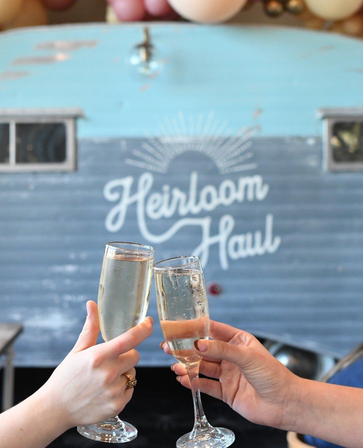 Two hands clink champagne flutes in front of a rustic trailer that has Heirloom Haul painted on the side.