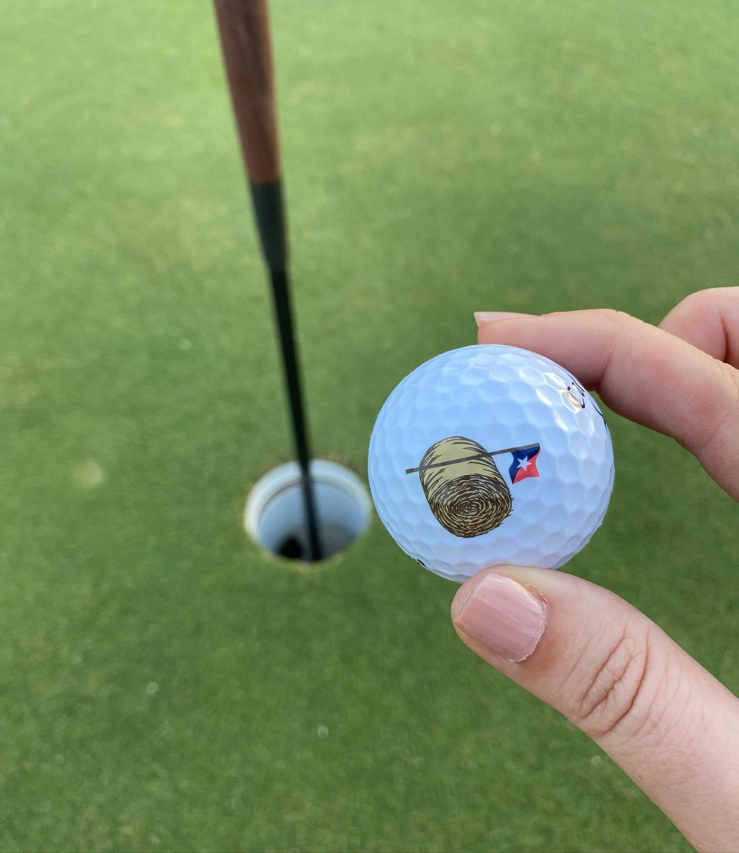 A hand holds up a branded golf ball with a golf hole in the background.