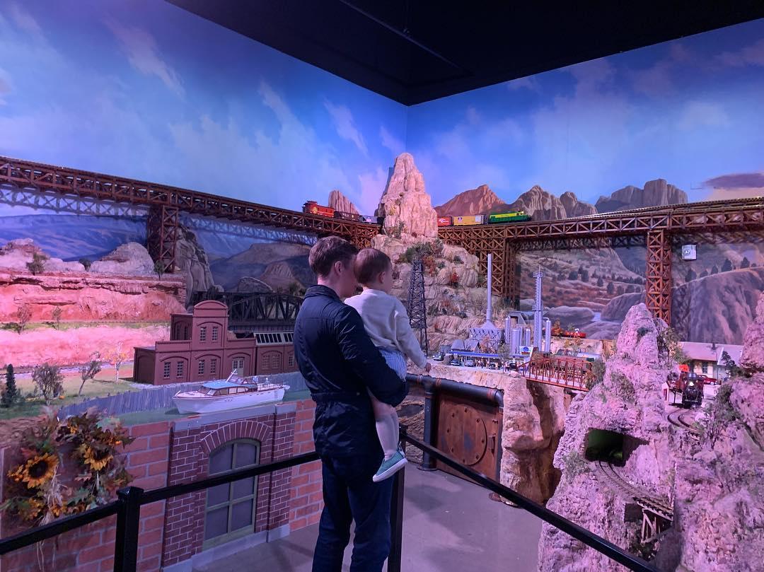 A father holds a toddler boy in his arms. The both are admiring a detailed miniature train set up.