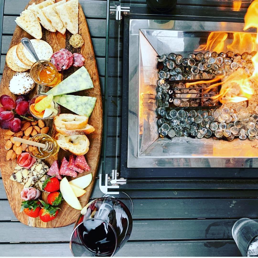 A charcuterie board of cured meats, assorted cheeses and baguette on a wooden plank next to a propane firepit.