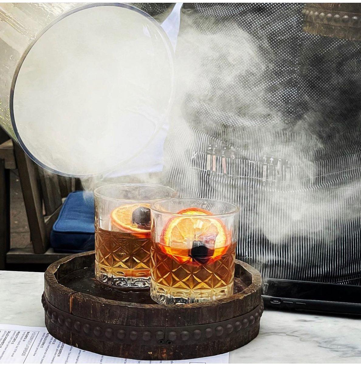 A glass dome is lifted off two craft cocktails with smoke billowing out from underneath.