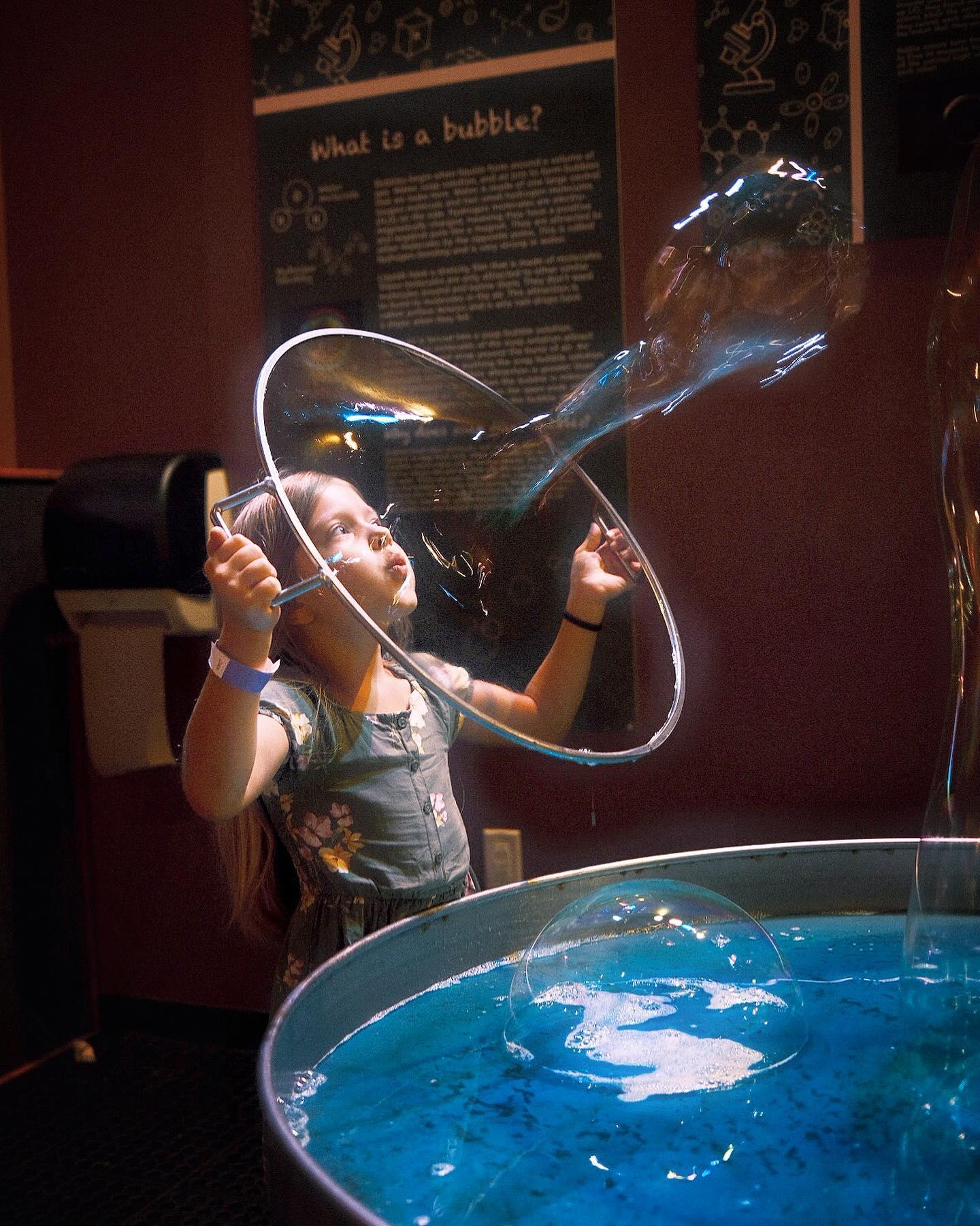 A little girl blows a large bubble through a soapy ring.