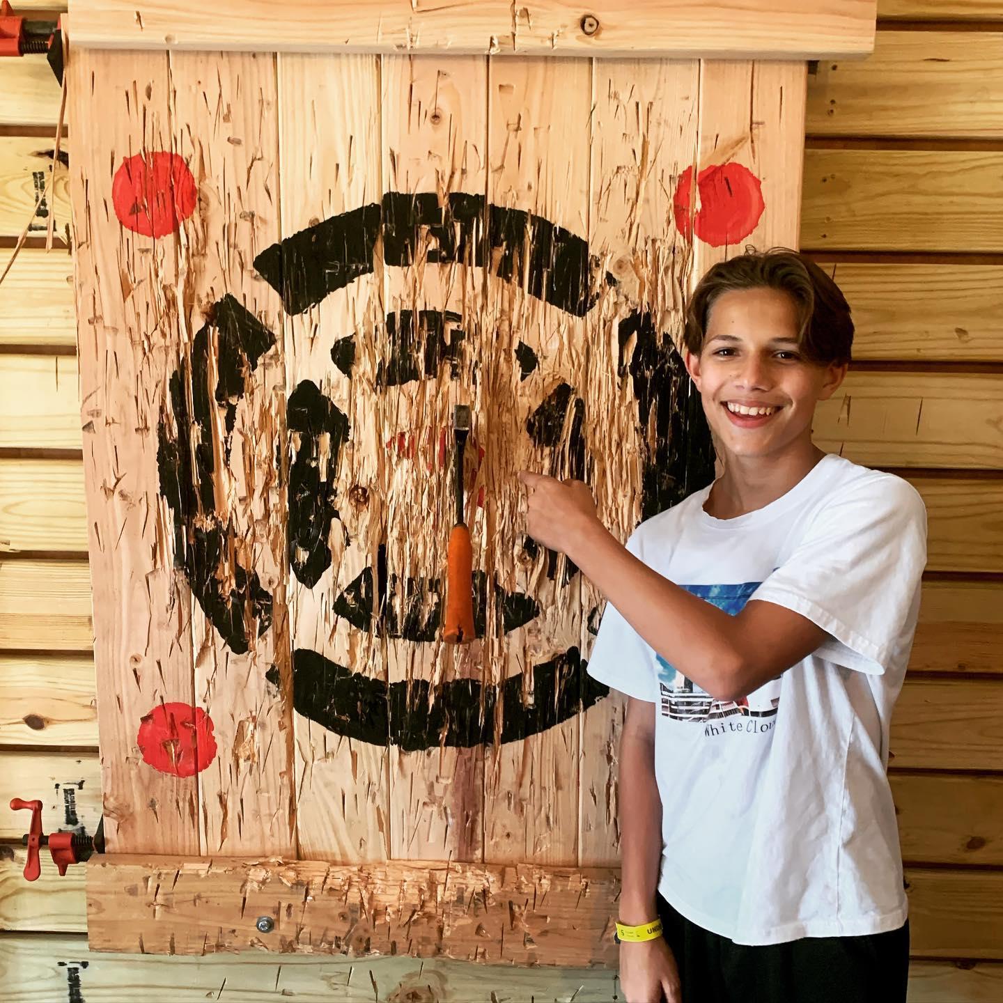 A boy poses next to an axe throwing wooden target with an axe.
