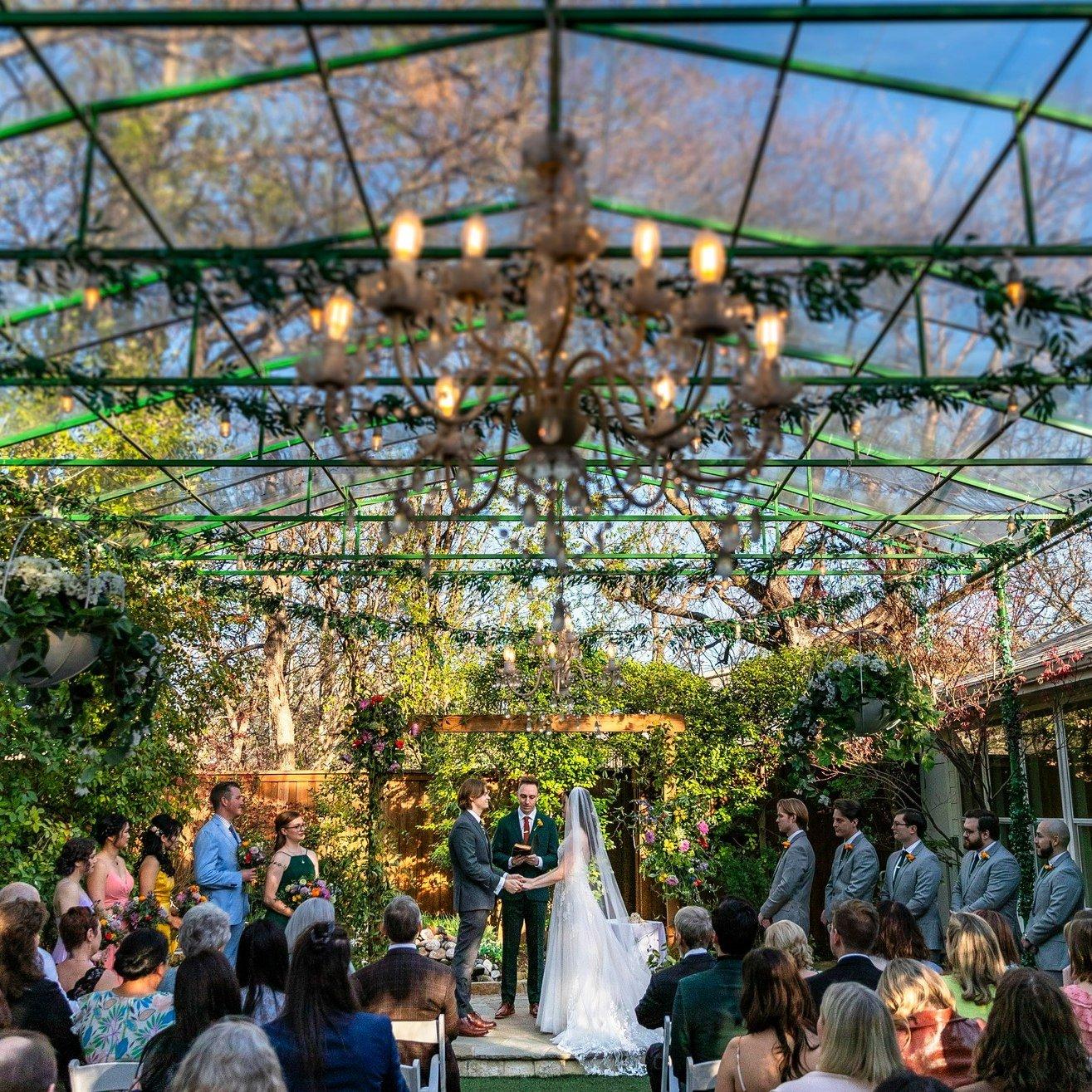 A colorful atrium with a grand chandelier. A bride and groom hold hands at the alter while guests watch.