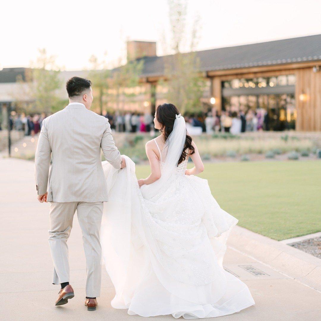 A bride and groom walk down a paved pathway to a venue in the distance at sunset.