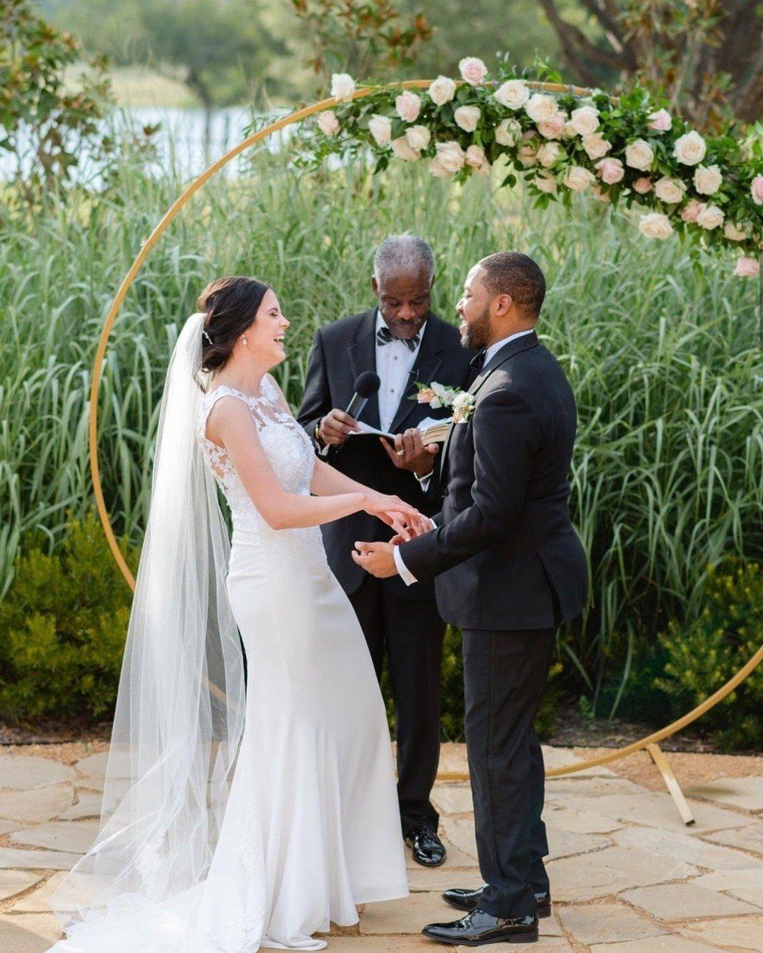 A bride and groom hold hands and laugh at the alter during their ceremony. The backdrop is lush greenery.