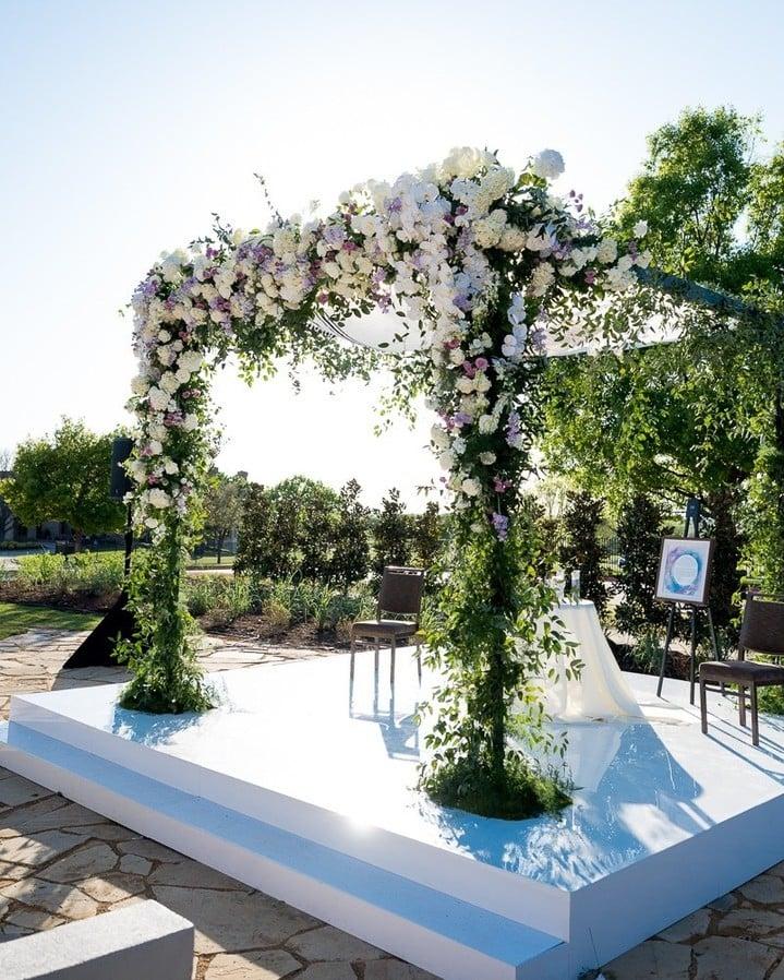 A floral arch made of greenery and white flowers at the alter of an outdoor wedding venue on a sunny day.