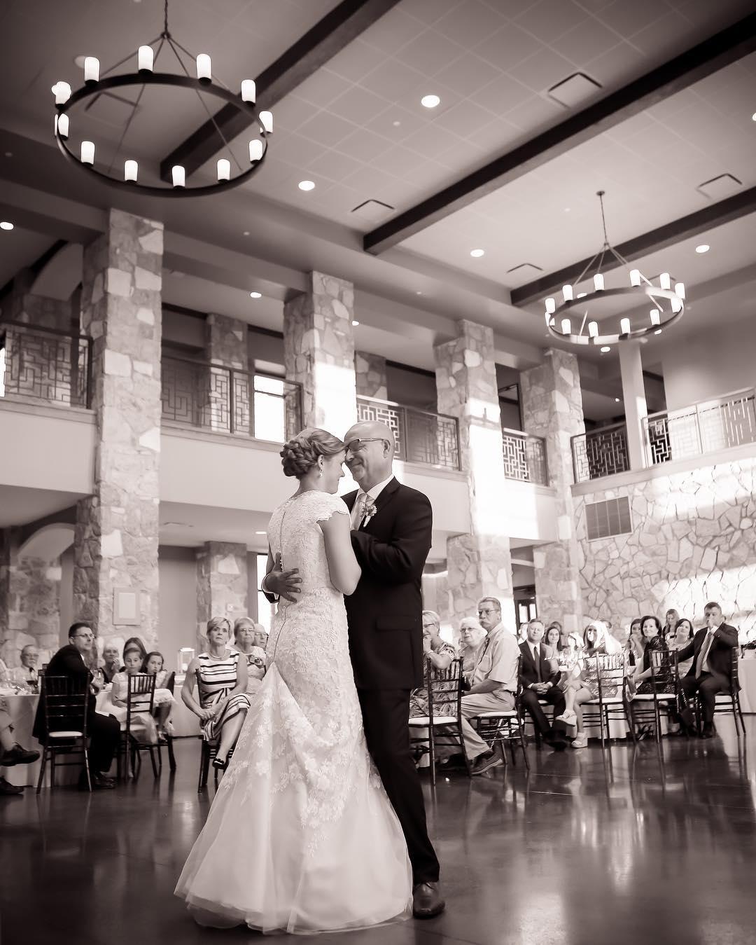 A black and white image of a bride and groom having their first dance in a ballroom while guests watch.