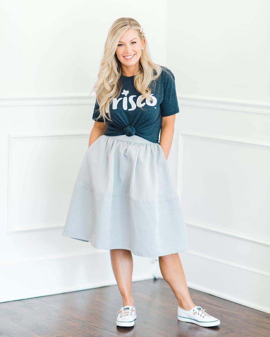 A woman with long, wavy, blond hair wearing a long white skirt and a blue t-shirt that says Frisco smiles for a photo with her hands in her pockets.