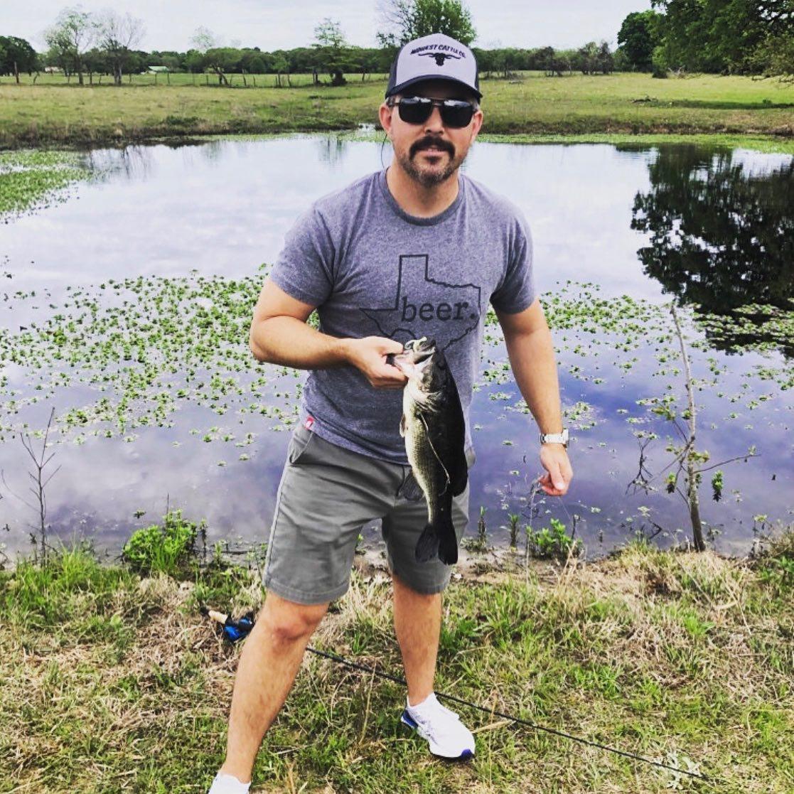 A man wearing a t-shirt that says Frisco poses while holding a fish he caught from the lake behind him.