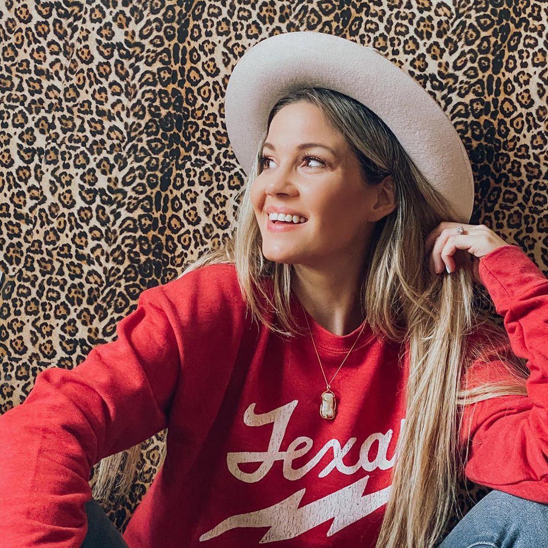 A blond woman wearing a red Frisco sweater and wide-brimmed hat poses for a photo in front of a leopard print backdrop.
