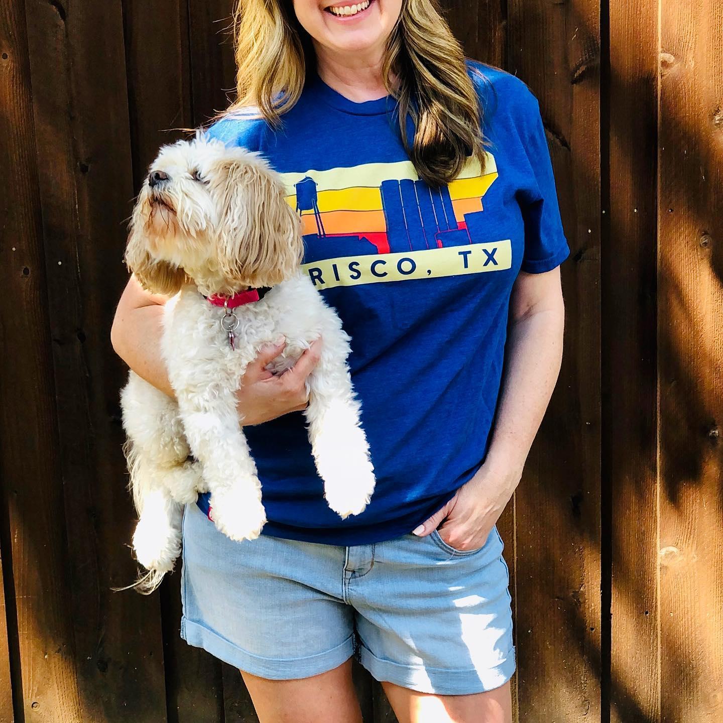 A woman wears a graphic t-shirt that says Frisco, Tx on it. She is holding a small dog and smiling for a photograph.