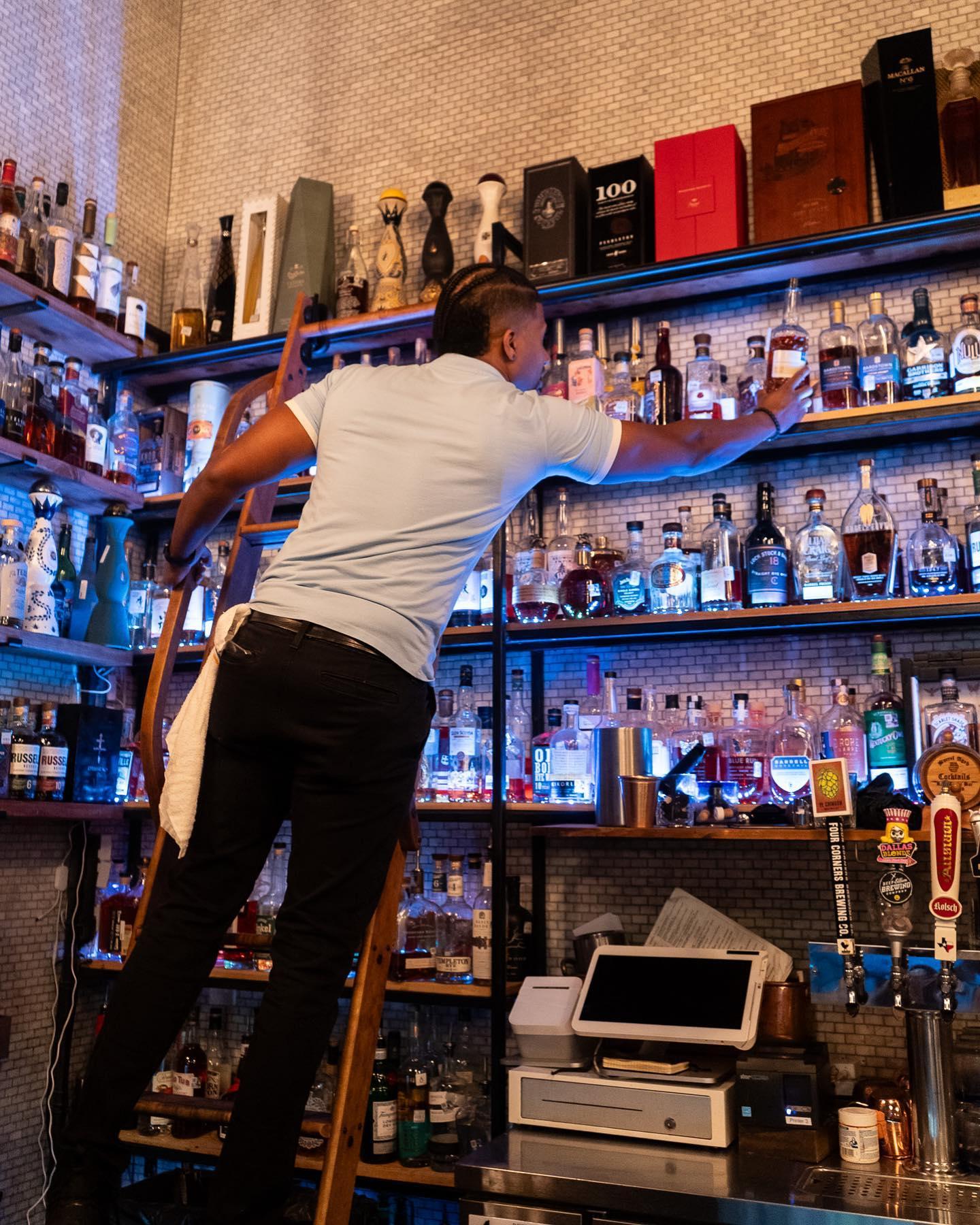a man on a ladder reaching to grab a bottle of alcohol
