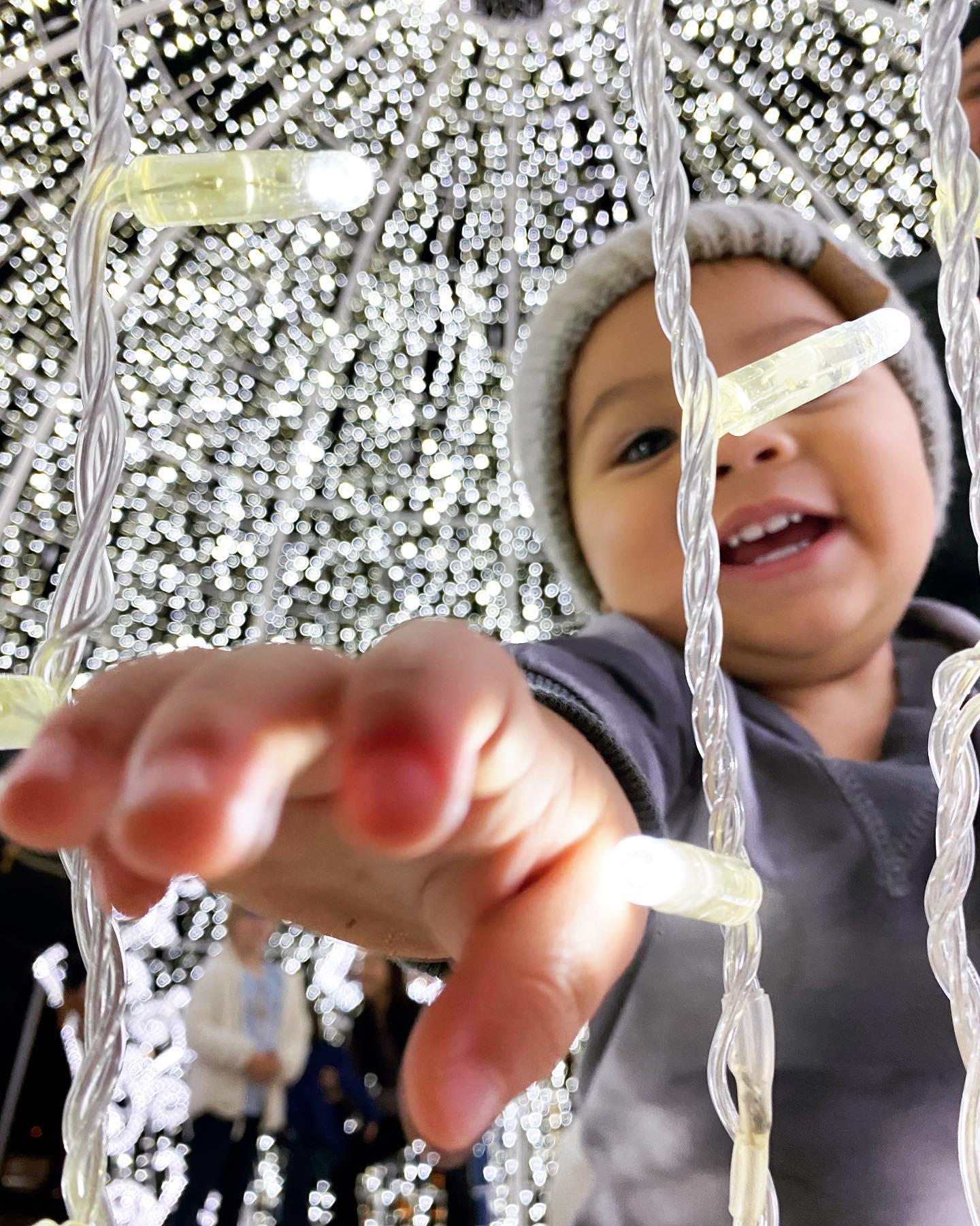 a little boy standing in a light sculpture reaching for a strand of lights
