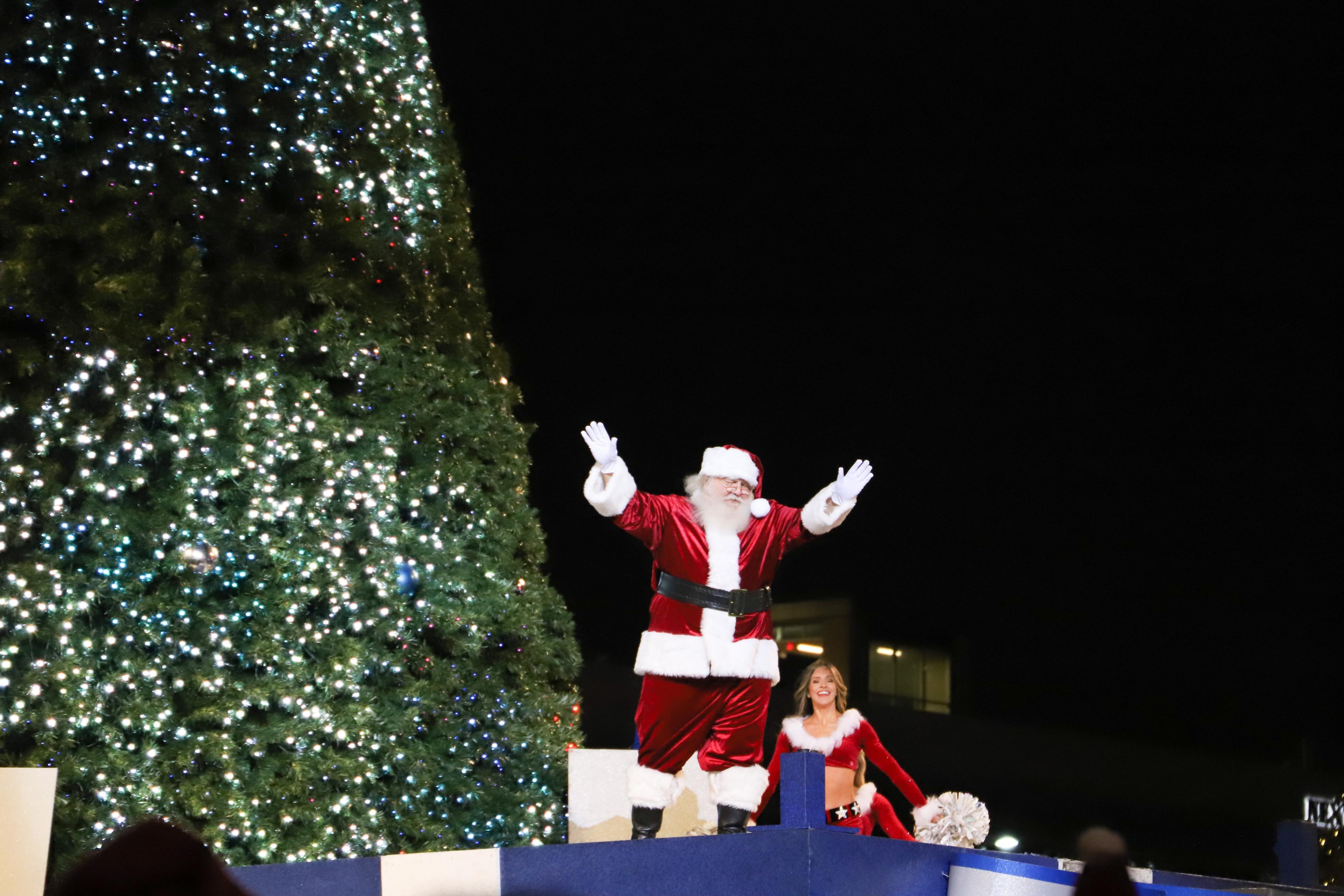 Santa raising his hands while standing next to a christmas tree