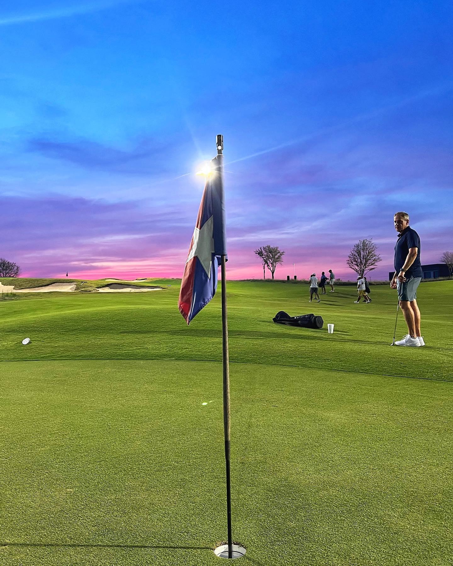 A man standing on the green next to the hole of a golf course with a beautiful blue and pink sunset in the background