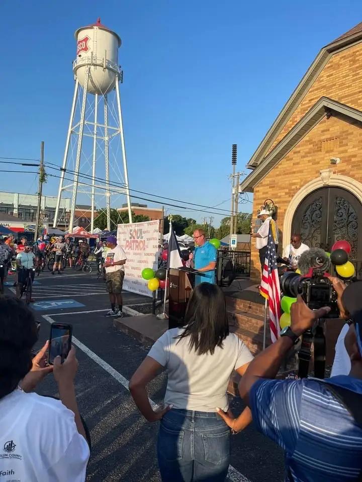 A large group of people gathered around a man speaking on a podium outside a church with a water tower in the distance