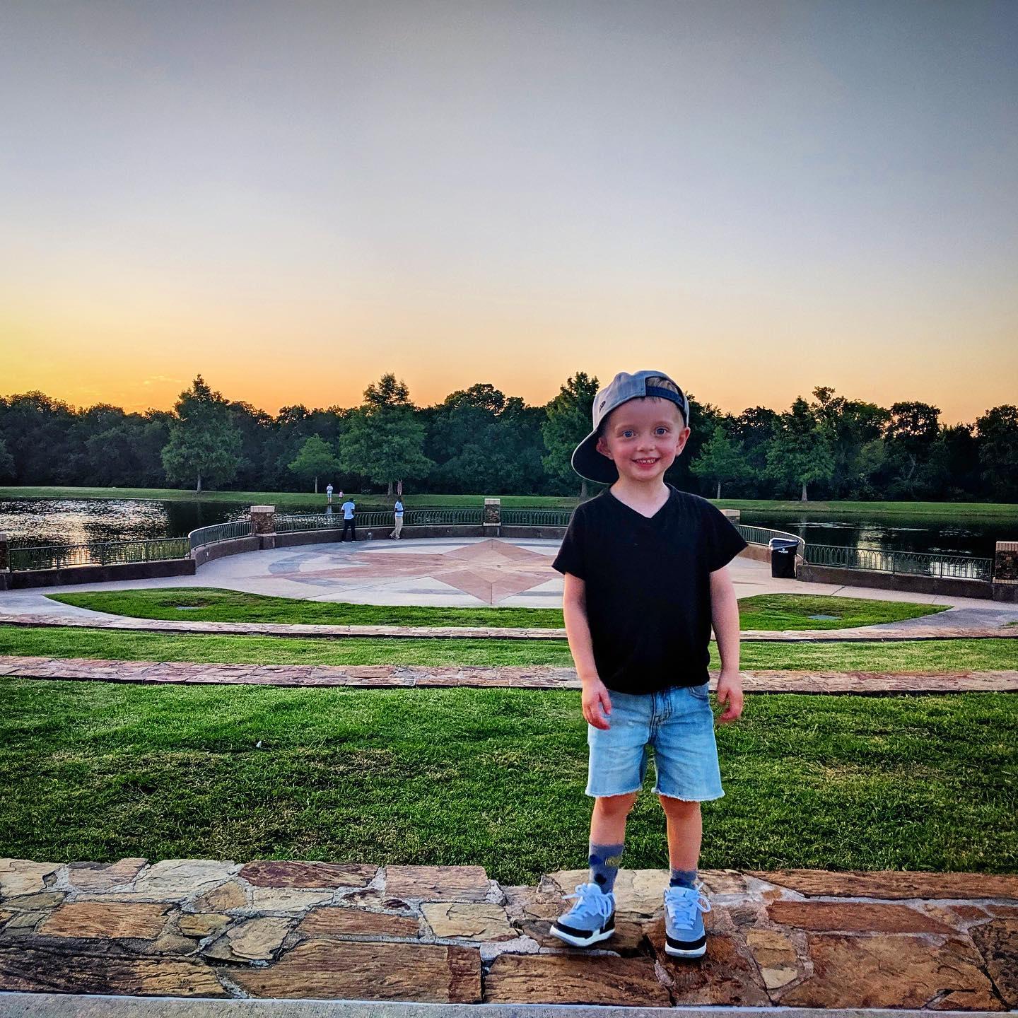 little boy in a baseball cap standing on a walkway with a lake behind him