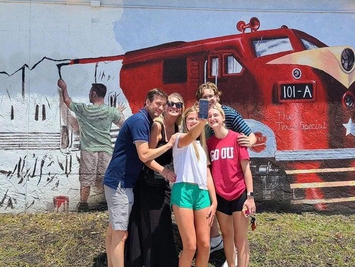 a family posing for a photo in front of a mural of a man painting a mural of a train